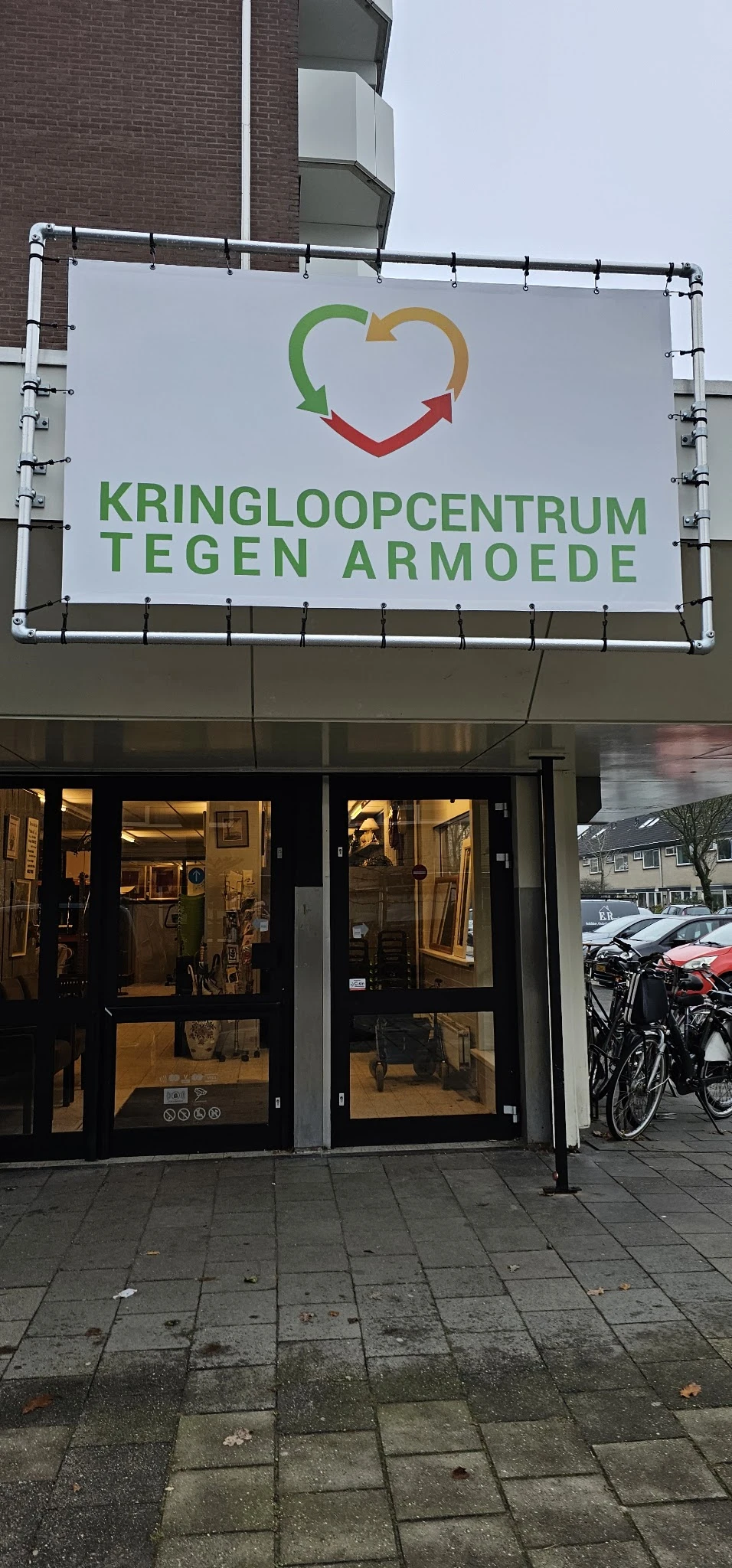 Winkelgevel van Kringloop Tegen Armoede met groene letters op wit bord, verlichte etalage met tweedehands spullen zichtbaar