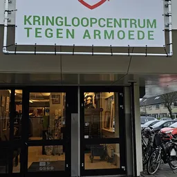 Winkelgevel van Kringloop Tegen Armoede met groene letters op wit bord, verlichte etalage met tweedehands spullen zichtbaar