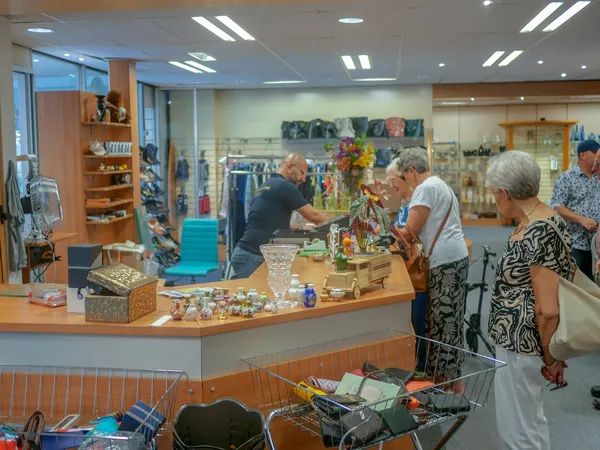 Winkelvloer van kringloopwinkel met klantenservice balie, klanten aan het winkelen, diverse spullen en rekken met kleding op 