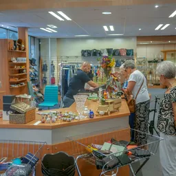 Winkelvloer van kringloopwinkel met klantenservice balie, klanten aan het winkelen, diverse spullen en rekken met kleding op 