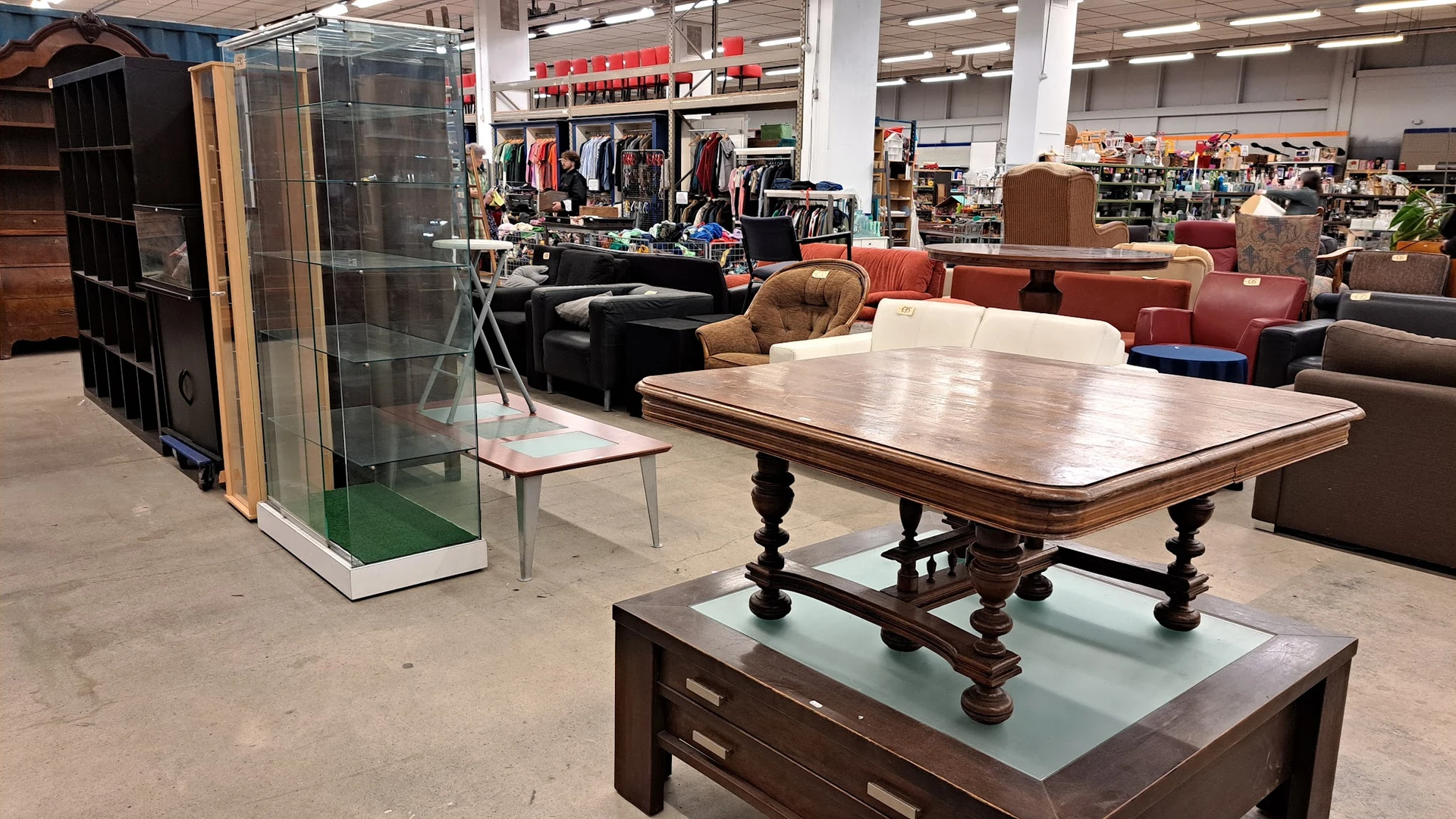 Winkelvloer met houten eettafel, bruine fauteuil, zwarte bank en displaykasten met kleding op achtergrond