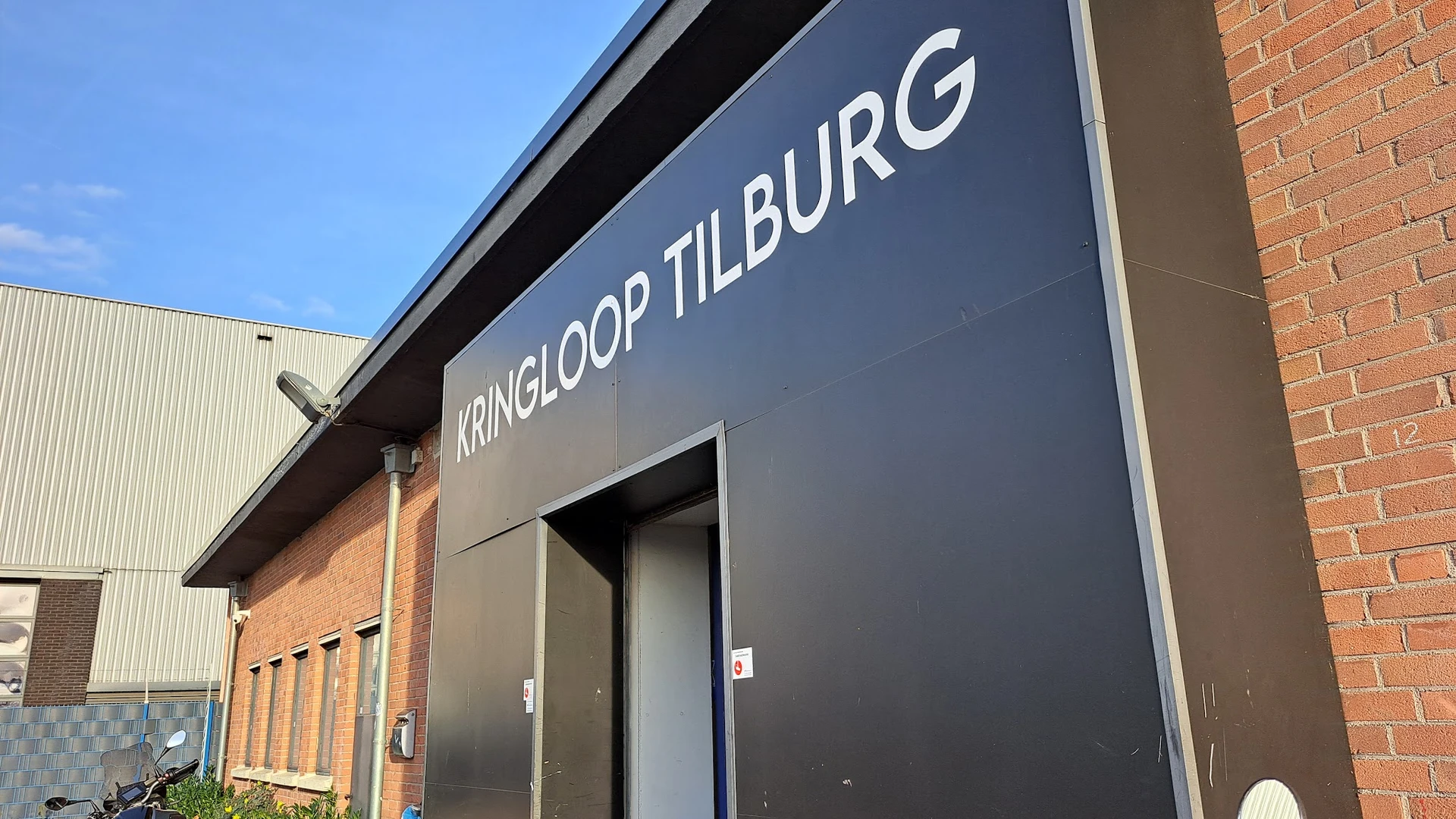 Grijze gevel van kringloopwinkel met wit uithangbord met winkelnaam en ingang