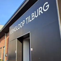 Grijze gevel van kringloopwinkel met wit uithangbord met winkelnaam en ingang