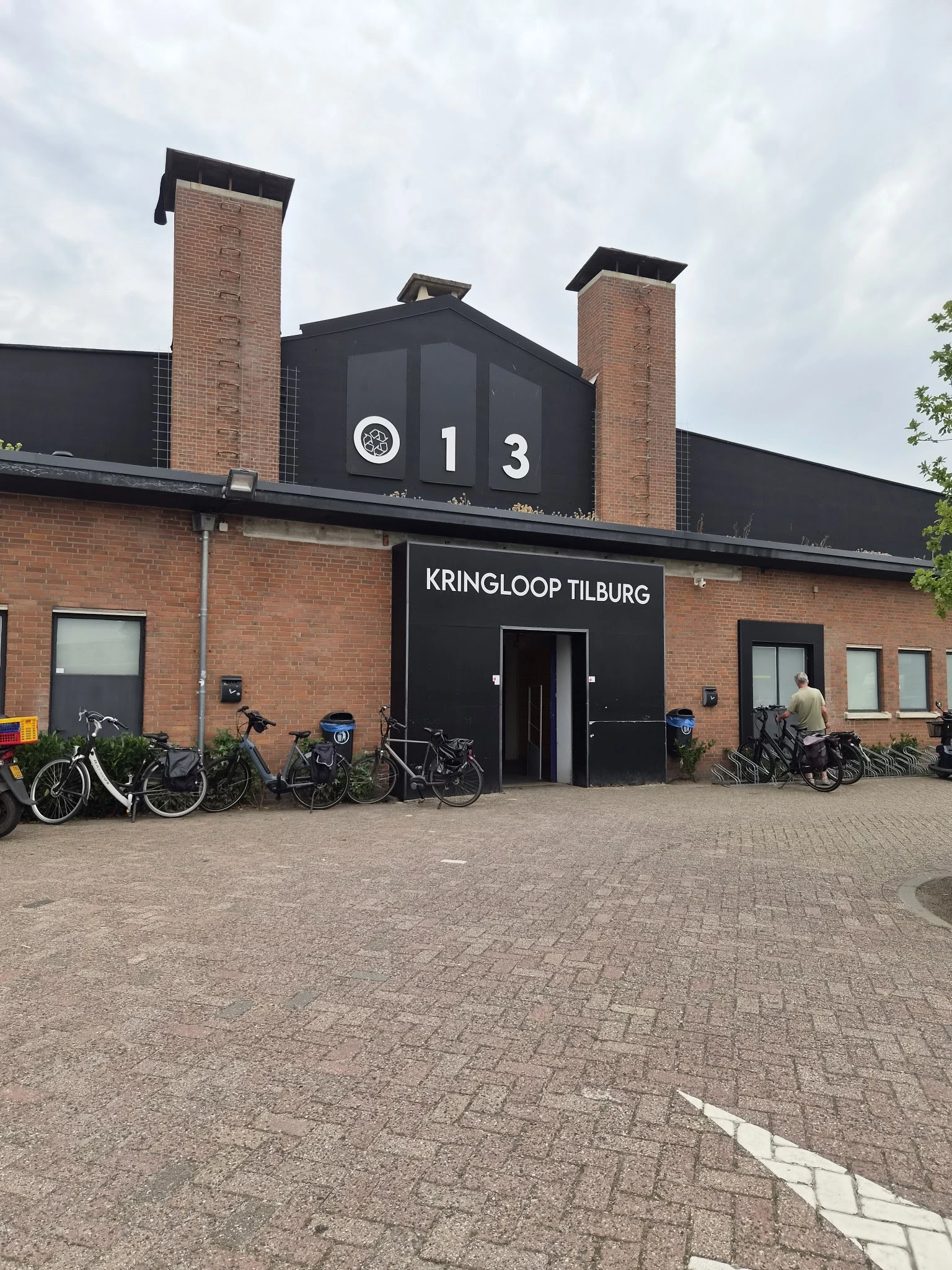 Gevel van kringloopwinkel met zwarte pui, bakstenen muren, twee schouwen en fietsen geparkeerd ervoor