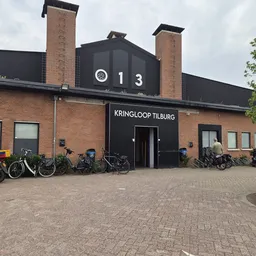 Gevel van kringloopwinkel met zwarte pui, bakstenen muren, twee schouwen en fietsen geparkeerd ervoor