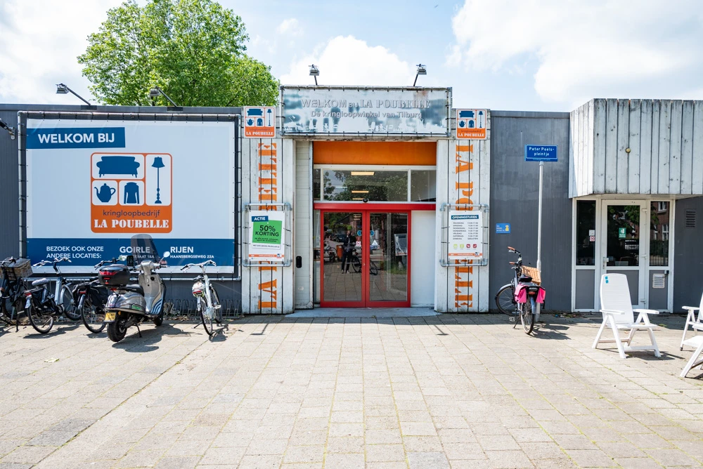 Gevel van kringloopwinkel met rode deuren, oranje details, fietsenstalling en welkomsbord aan de zijkant