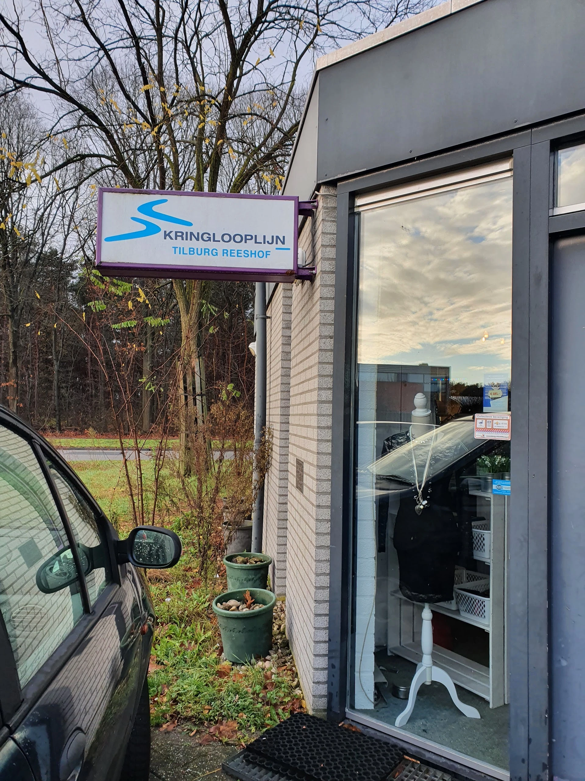 Gevel van Kringlooplijn Tilburg Reeshof met blauwe grijze winkelruit en uithangbord, zijkant auto zichtbaar.