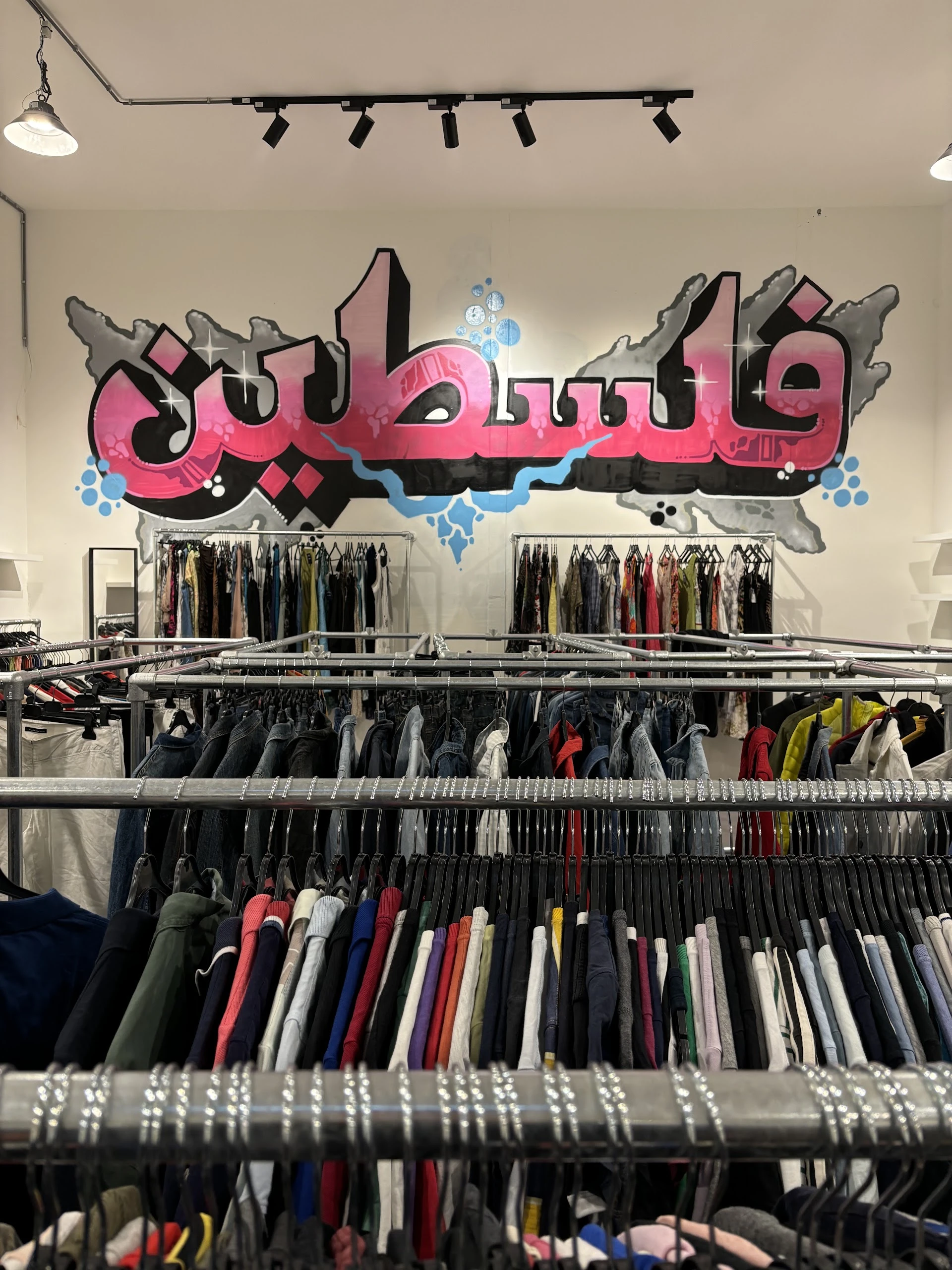 Rekken met tweedehands kleding in verschillende kleuren, onder een roze neonlogo aan de muur