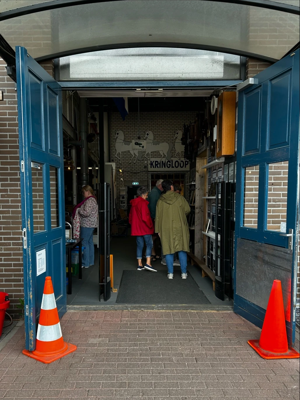 Bezoekers lopen door een kringloopwinkel met blauwe deuren, stellingen met spullen en een 'KRINGLOOP' bord aan de muur