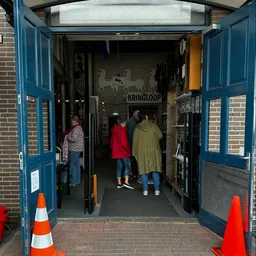 Bezoekers lopen door een kringloopwinkel met blauwe deuren, stellingen met spullen en een 'KRINGLOOP' bord aan de muur