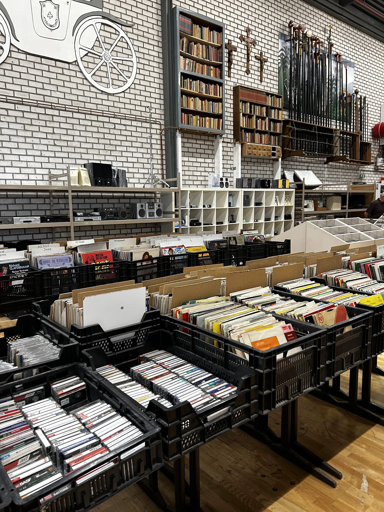 Zwarte kunststof kratten met cd's, dvd's en boeken op een tafel in de kringloopwinkel, met stellingen op de achtergrond