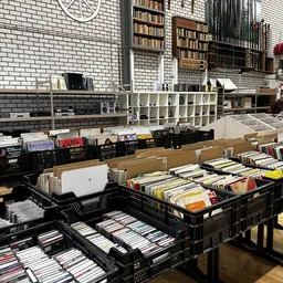 Zwarte kunststof kratten met cd's, dvd's en boeken op een tafel in de kringloopwinkel, met stellingen op de achtergrond