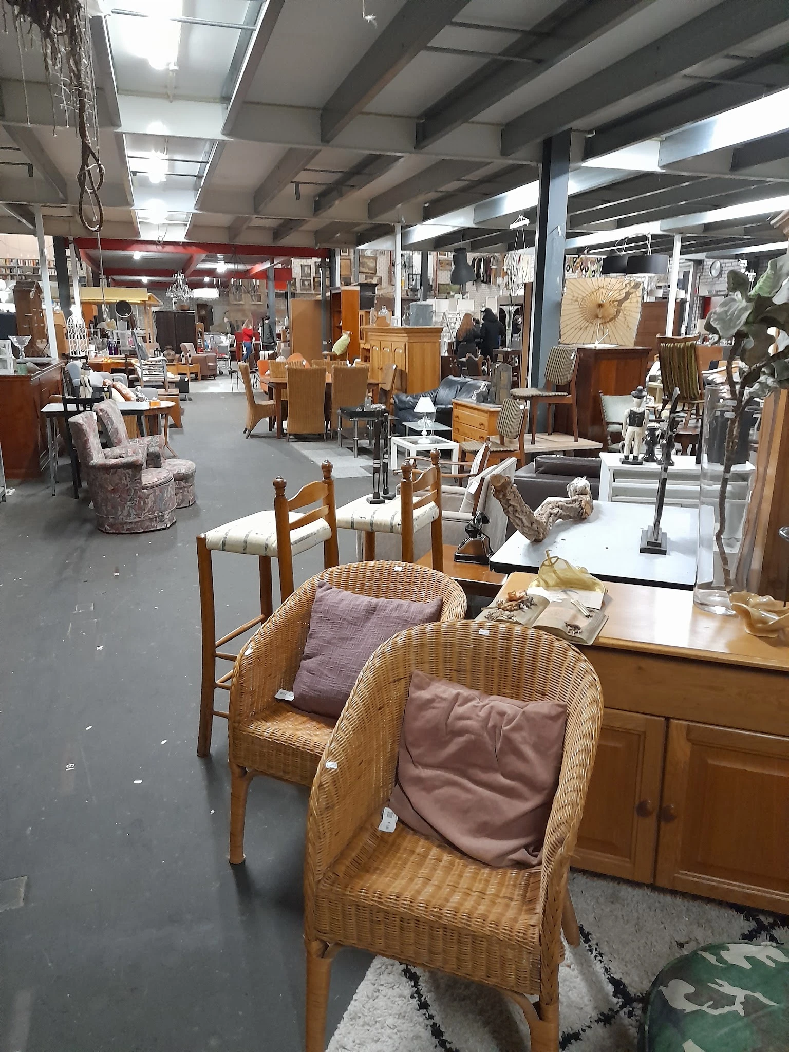 Winkelvloer van kringloopwinkel met rieten stoelen, houten tafels en diverse meubels op de achtergrond