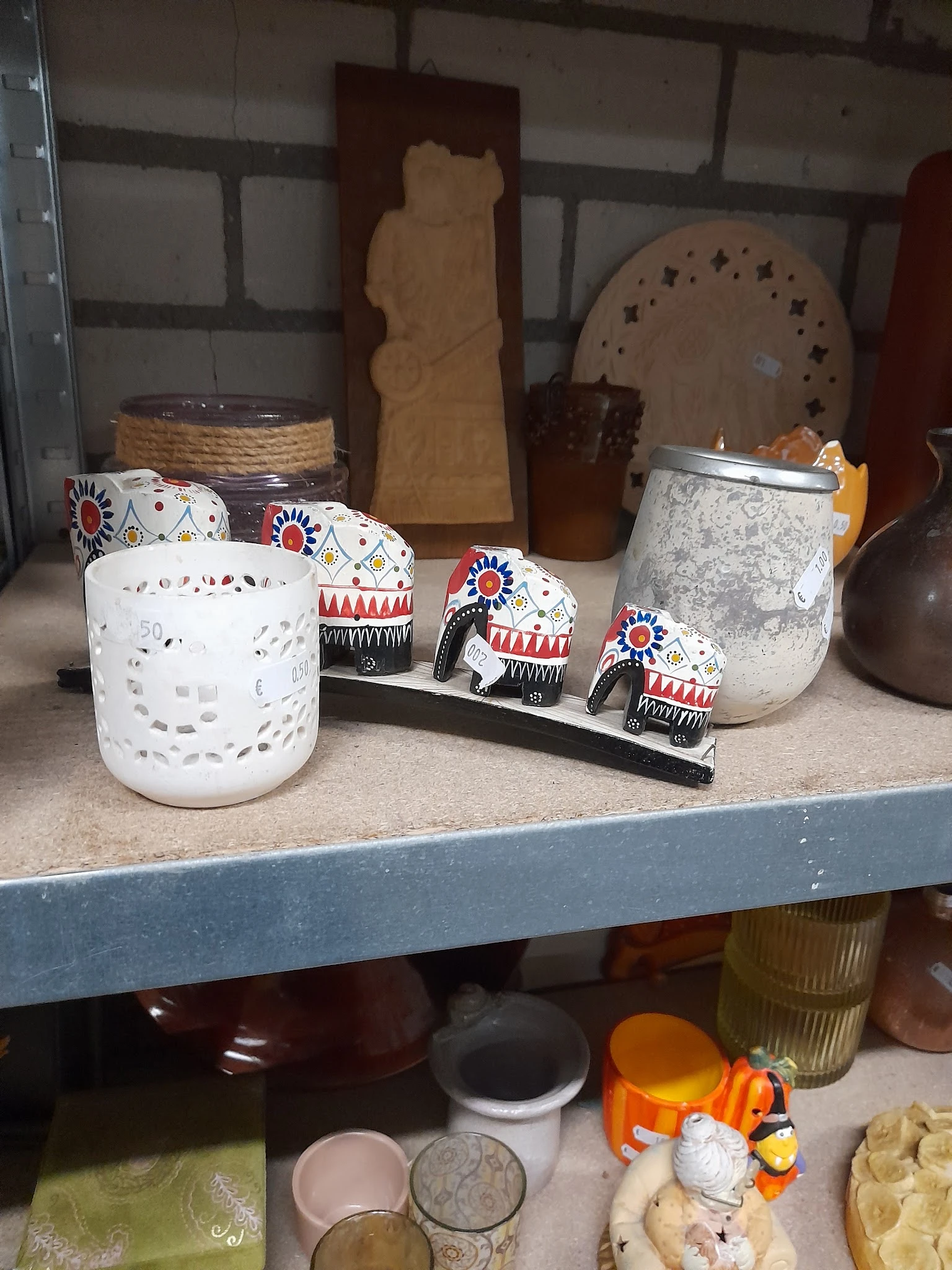 Uitgestalde decoratieve items op een plank: witte kandelaar, grijze pot en drie beschilderde olifantjes met etnische patronen
