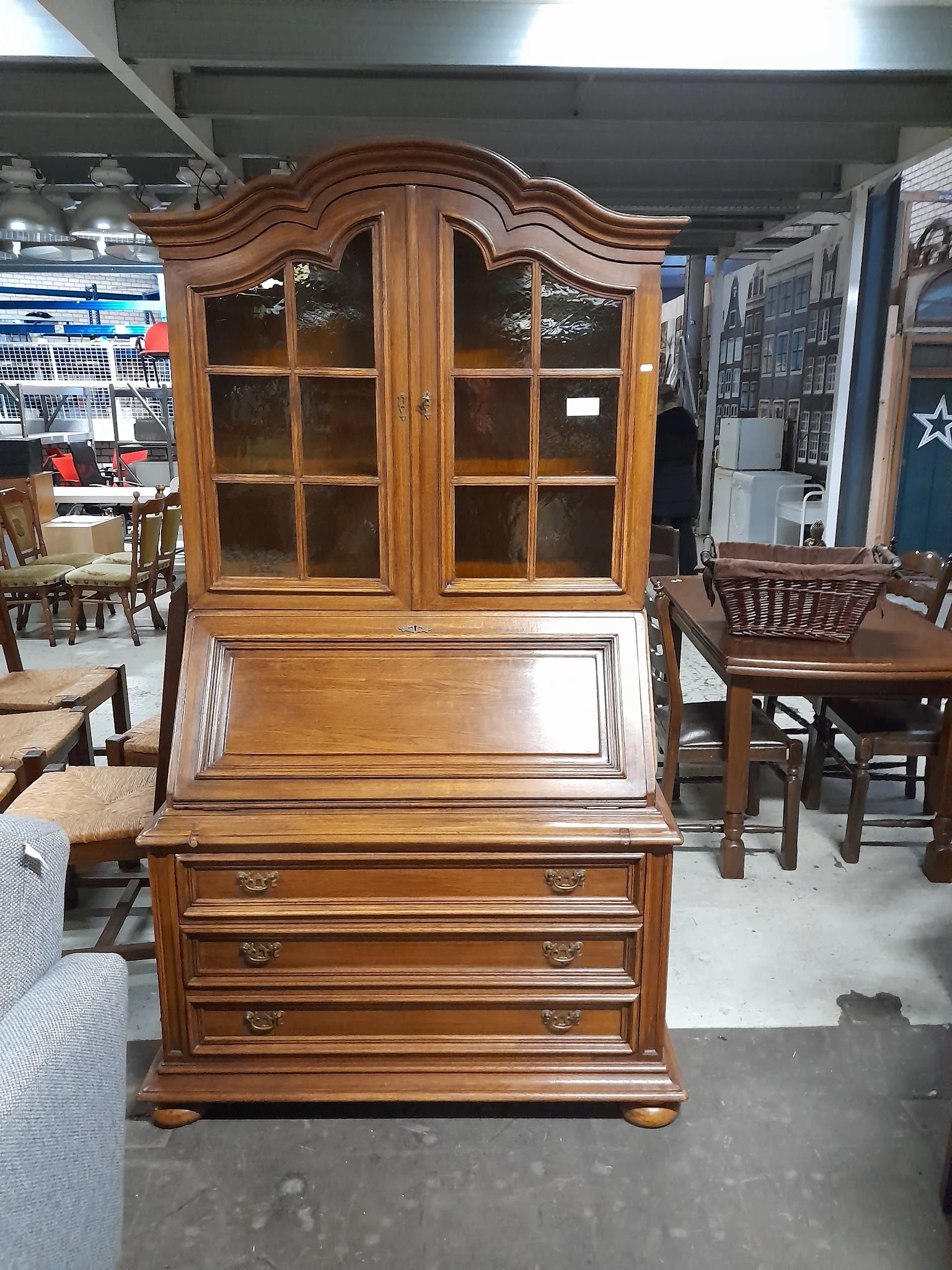 Houten secretaire met glazen kastdeuren bovenop en twee laden, in een kringloopwinkel tussen andere meubels