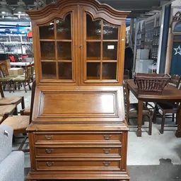 Houten secretaire met glazen kastdeuren bovenop en twee laden, in een kringloopwinkel tussen andere meubels