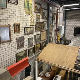 Muur met uitgestalde schilderijen en kunstwerken in houten en donkere lijsten, werktafel op voorgrond