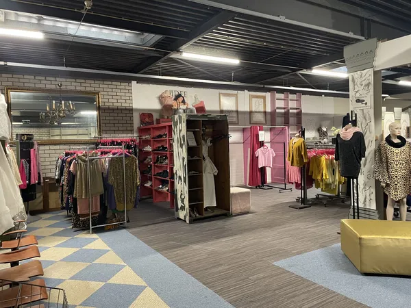 Winkelinterieur met rekken vol dameskleding in verschillende kleuren, rode stellingen, kleedkamers en gele poef op grijze vlo