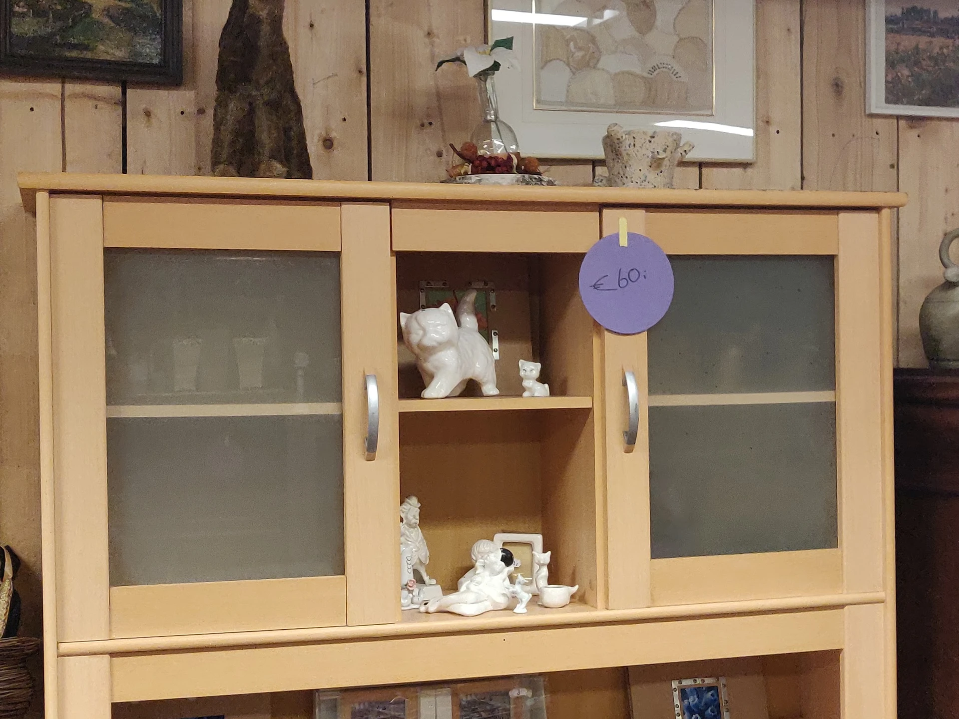 Houten vitrinekast met glazen deuren, gevuld met witte decoratieve objecten en serviesgoed, met een paars prijskaartje van €6