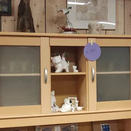 Houten vitrinekast met glazen deuren, gevuld met witte decoratieve objecten en serviesgoed, met een paars prijskaartje van €6