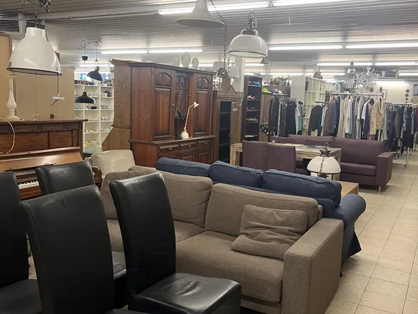 Winkelvloer met diverse meubels: grijsbeige bank, zwarte stoelen, houten kastmeubel, en kleding op rekken op de achtergrond