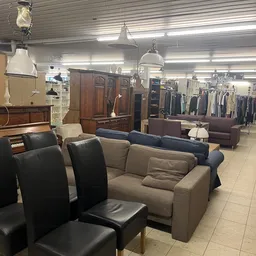 Winkelvloer met diverse meubels: grijsbeige bank, zwarte stoelen, houten kastmeubel, en kleding op rekken op de achtergrond