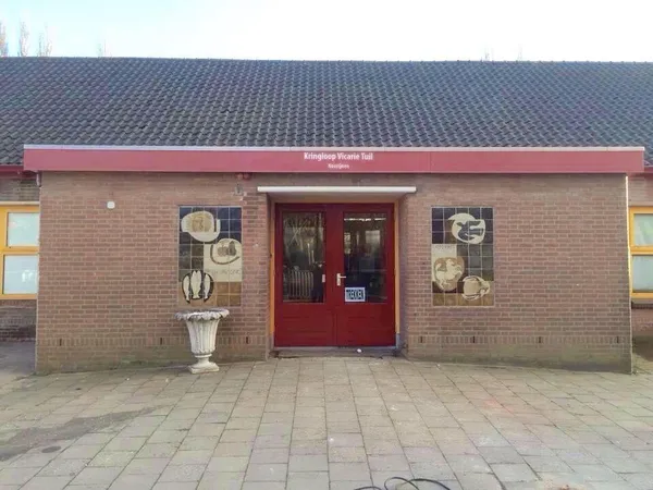 Baksteinen gevel met rode deur en luifel van kringloopwinkel, decoratieve bordjes aan de muur