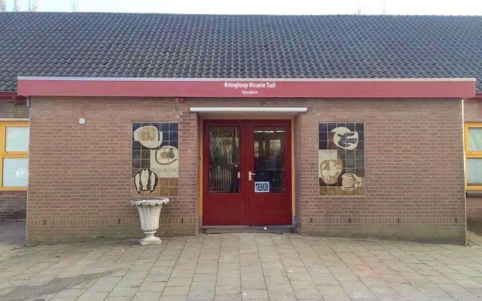 Bakstenen gevel van kringloopwinkel met rode deuren en rode dakrand, ingangsdeur met ramen, decoratieve bordjes aan wand