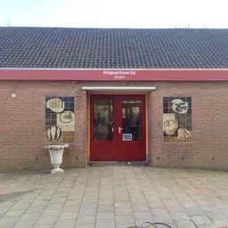 Baksteinen gevel met rode deur en luifel van kringloopwinkel, decoratieve bordjes aan de muur