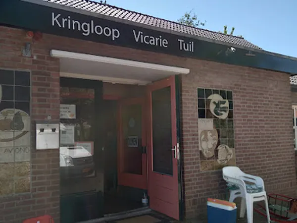 Voorgevel van kringloopwinkel met rode deur, bakstenen muur en winkelnaam op bord, met enkele uitgestalde items buiten