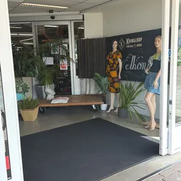 Winkelinterieur met etalagepop in gele jurk, houten bank, potplanten en zwarte achtergrondwand met opschrift.