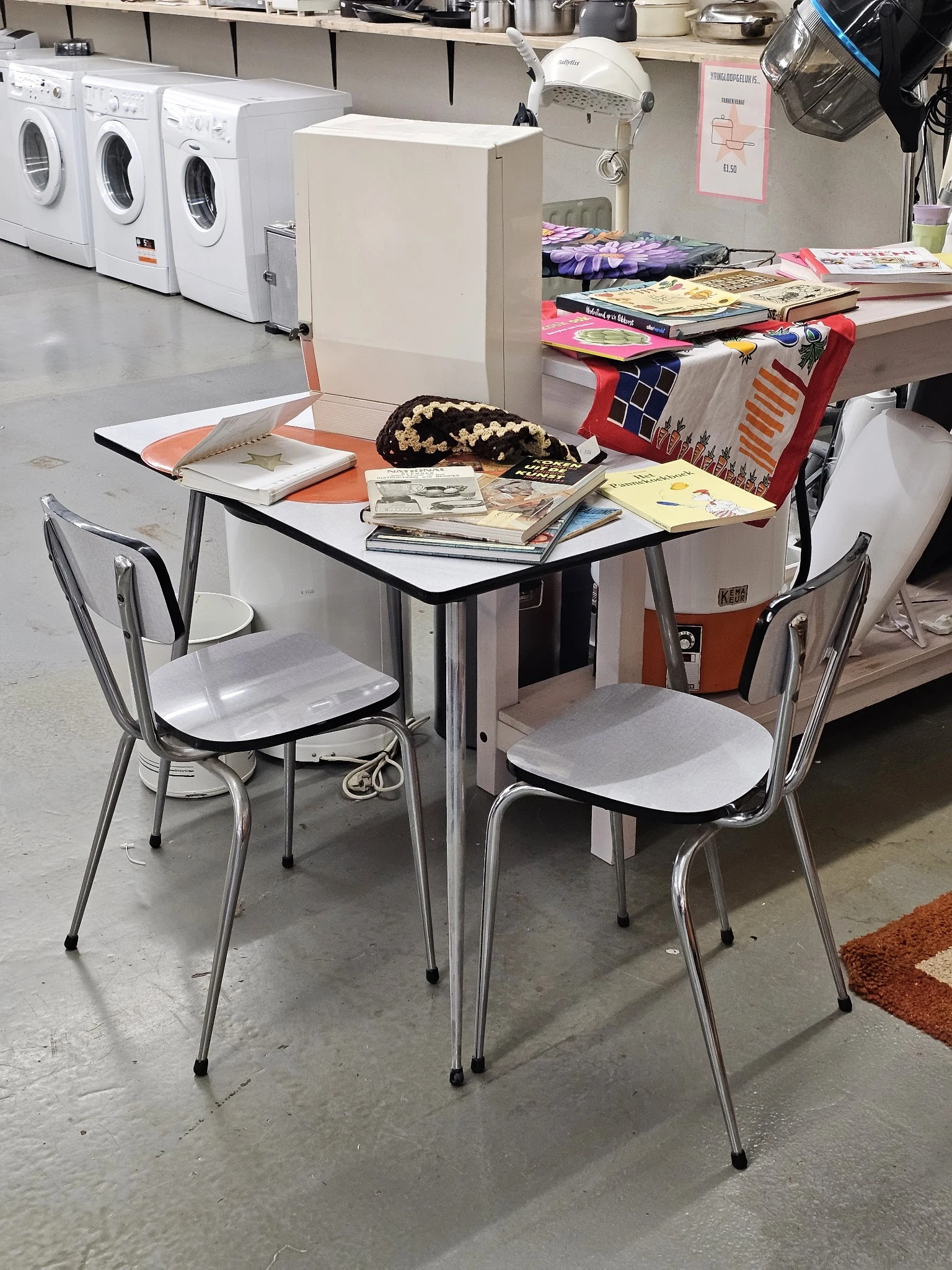 Eettafel met twee stoelen en stapels spullen op de tafel en op een rood rek op de achtergrond