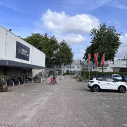 Voorkant van kringloopwinkel met witte gevel, ingang met fietsen geparkeerd, parkeerplaats met auto's en rode vlaggen