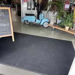 Winkelinterieur met donkergrijs tapijt, zwarte deurmat, houten plank met planten en een blauwe vintage scooter op betonnen vl