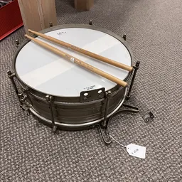 Zwarte snaredrum met witte vel en houten drumstokken op grijze vloer