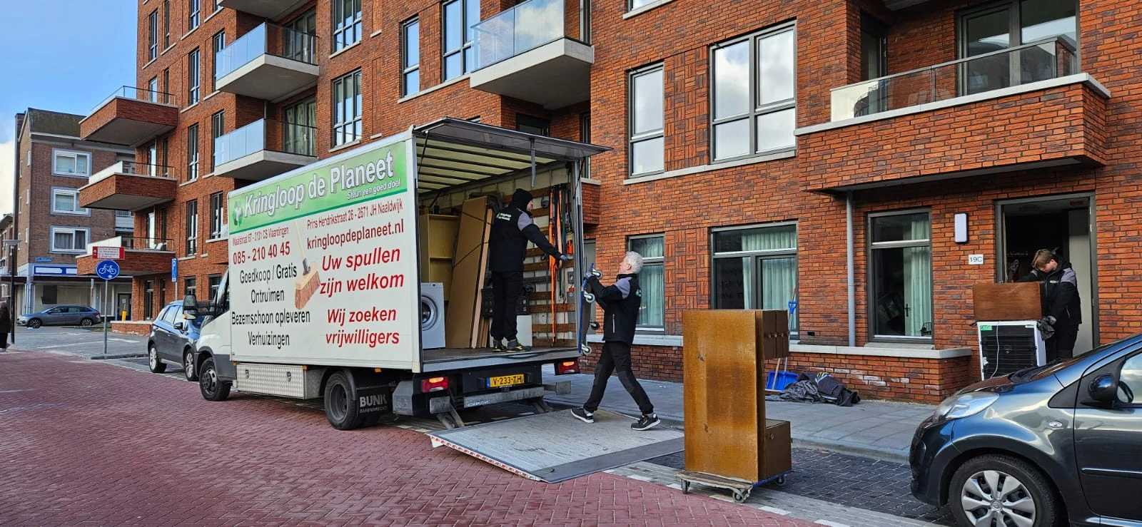 Medewerkers laden spullen uit een bedrijfsbus van Kringloop Planeet bij een bakstenen gebouw