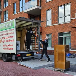 Medewerkers laden spullen uit een bedrijfsbus van Kringloop Planeet bij een bakstenen gebouw