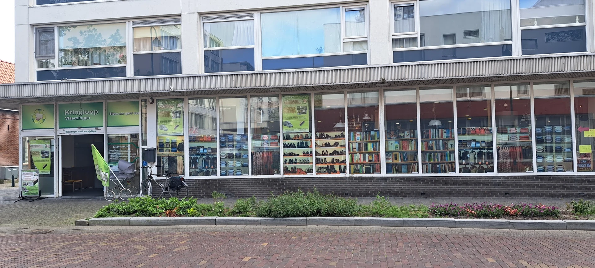 Winkelgevel met grote ramen waarin boekenkasten en vintage spullen zichtbaar zijn, met groene planten in bakken ervoor
