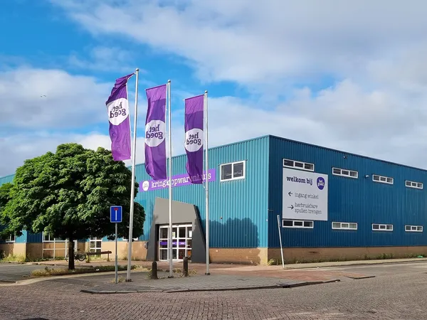 Gevel van een kringloopwinkel met groene metalen constructie, paarse banners en een tuinboom ervoor