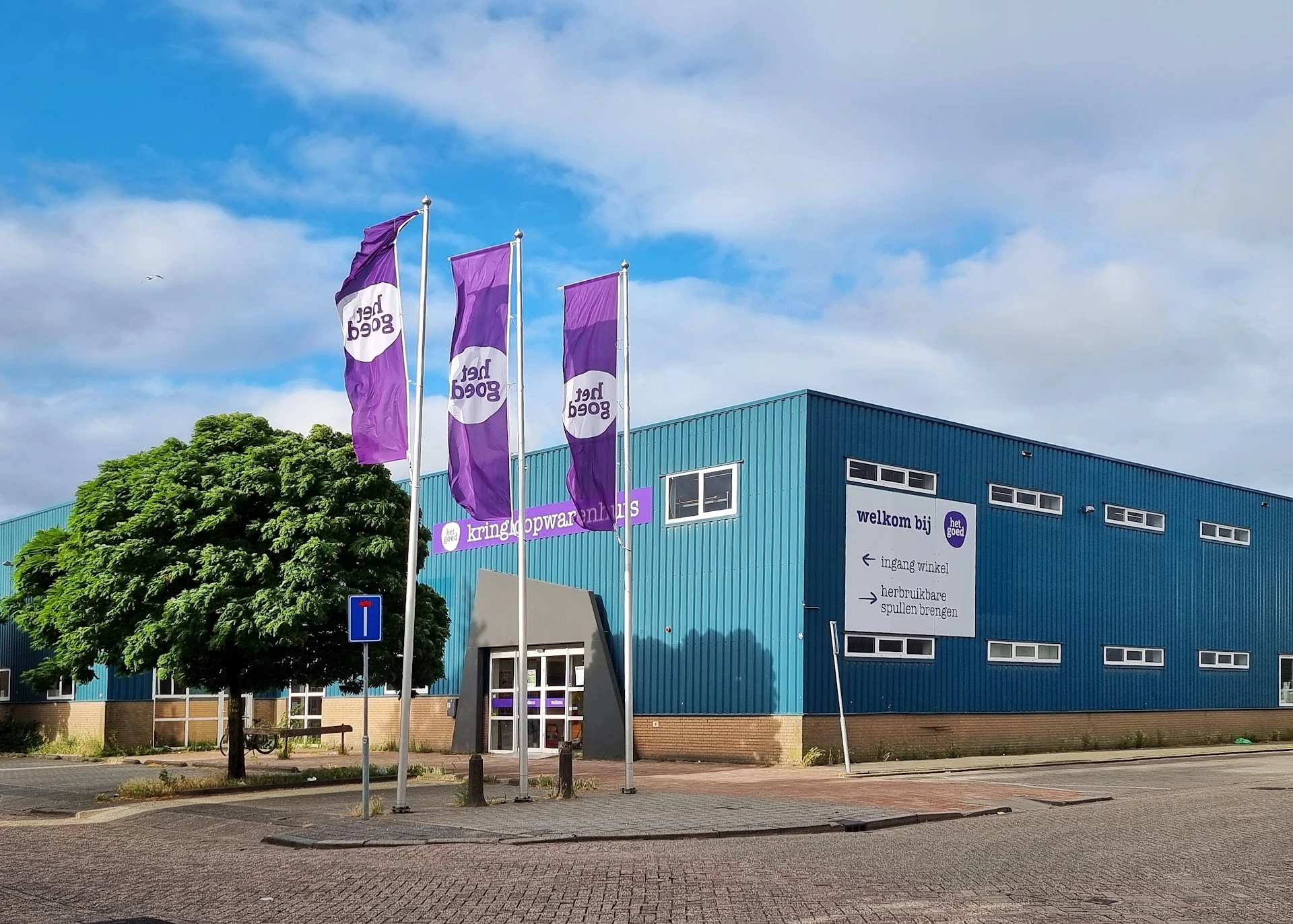 Gevel van een kringloopwinkel met groene metalen constructie, paarse banners en een tuinboom ervoor