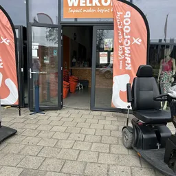 Ingang van kringloopwinkel met oranje banner en zwarte rolstoel op terras