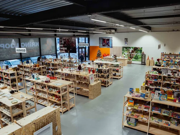 Overzicht van winkelvloer met houten stellingen vol spullen, open ruimte, ramen en oranje accenten aan achterwand