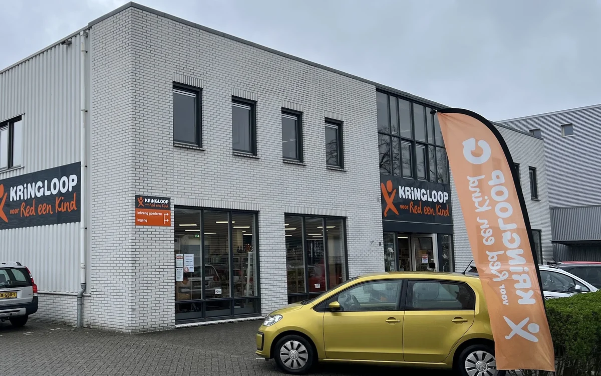 Witte bakstenen gevel van kringloopwinkel met grote ramen en oranje banner, gele auto geparkeerd ervoor