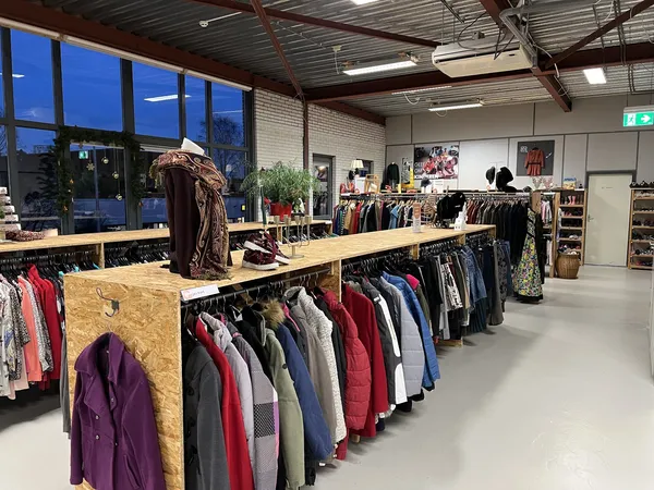 Ruime winkelruimte met houten displaytafels vol kleding, stellingen met kleding tegen de muren, en grote ramen met blauwe avo