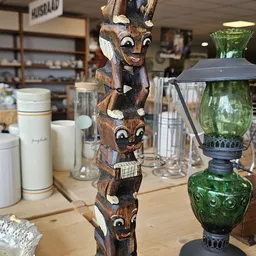 Houten beeldhouwwerk met gestapelde gezichten naast huishoudelijke items en glazen karaffen in de winkel
