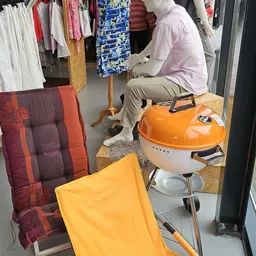Uitgestalde gebruiksartikelen in de kringloopwinkel: oranje barbecue, geel en bordeaux stoelen, en diverse kleding op achterg
