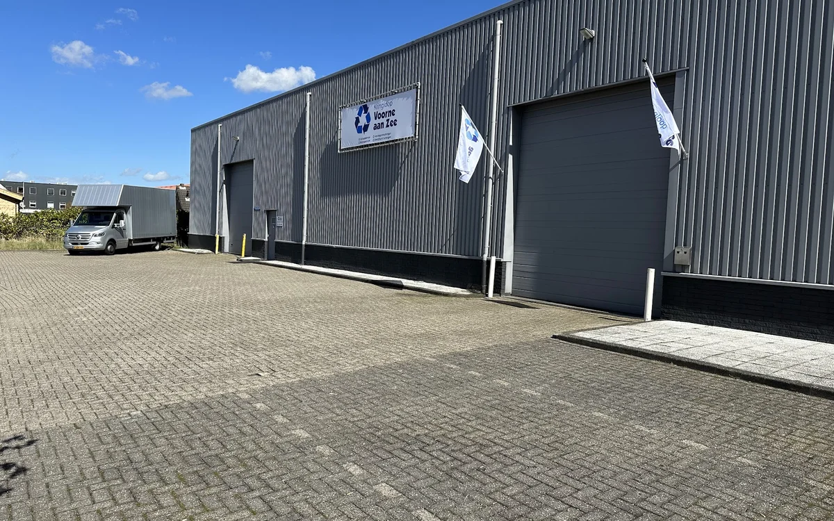 Grijze bedrijfshal met twee grote donkergrijze deuren en wit logo bord, parkeerterrein met tegels