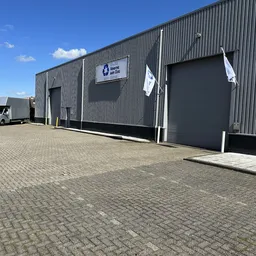 Grijze bedrijfshal met twee grote donkergrijze deuren en wit logo bord, parkeerterrein met tegels