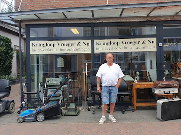 Voorgevel van kringloopwinkel met glazen pui, eigenaar staande voor ingang, uitstallingen met gebruiksgoederen zichtbaar