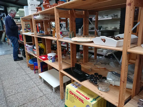 Houten stellingen met diverse spullen en voorwerpen in een kringloopwinkel, met een persoon op de achtergrond